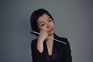 A headshot of Moyue Zhou, holding her conducting baton under her face and looking at the camera